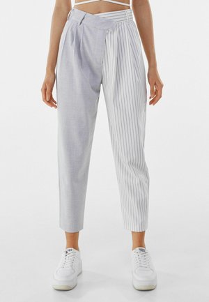 Vrouw draagt een broek met hoge taille, het ene been in grijs ruitpatroon en het andere in witte krijtstreep, gecombineerd met witte sneakers.