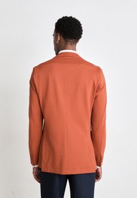 Orangemelerad blazer med notch-krage, enkel ryggslits och knappdetaljer vid ärmsluten. Stylist med en vit undertröja.