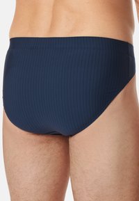 Marineblaue Schwimmslips mit einem Nadelstreifen-Muster, elastischem Bund und minimalistischem Design, die die Rückansicht auf heller Haut zeigen.