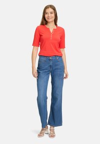 Rød kortærmet henley skjorte med knaplukning sat sammen med lyseblå bredbenede jeans. Model iført hvide stroppesandaler. Enkel baggrund.