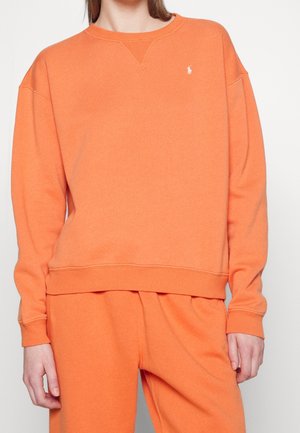 Personne portant un ensemble assorti sweat-shirt et pantalon orange, avec un petit logo blanc Polo Ralph Lauren sur la poitrine gauche du sweat-shirt.