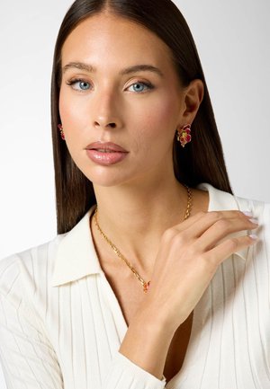 Woman with blue eyes and straight brown hair wearing red and gold floral earrings and matching necklace, dressed in a white ribbed top.