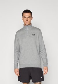 Šedý pulover na čtvrt zip v měkkém materiálu, s dlouhými rukávy, žebrovanými manžetami a černým logem Puma na hrudi. Doplněno černými šortkami.