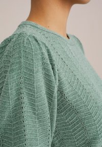 Pulover verde texturat cu un model cu nervuri și detalii subtile de ochiuri. Dispune de un gât rotund și mâneci scurte, plisate.