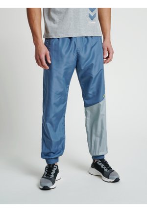 Mężczyzna noszący błyszczące niebieskie spodnie dresowe z jasnoszarys łatą na jednej nodze, szary T-shirt i czarno-szare trampki stojący na białej podłodze.