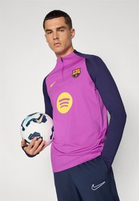 Camisa do FC Barcelona em rosa com mangas azul-marinho, apresentando um logótipo e um gráfico amarelo do Spotify. Um jogador segura uma bola de futebol com padrão.