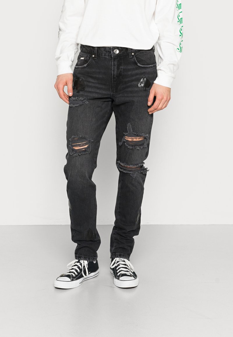 Black denim jeans with a slim fit, distressed detailing including ripped knees, and subtle fabric patches. Styled with black sneakers.