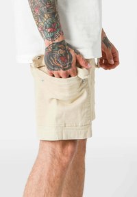 Pantaloni corti beige con una vestibilità rilassata, dotati di una tasca laterale e chiusure a scatto. Il tessuto è leggero e morbido, offrendo un aspetto casual.
