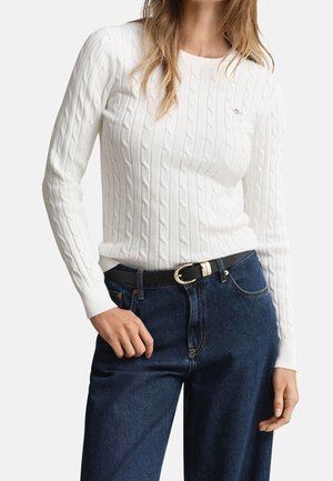 Femme portant un pull blanc en maille torsadée rentré dans un jean bleu foncé avec une ceinture noire à boucle dorée.