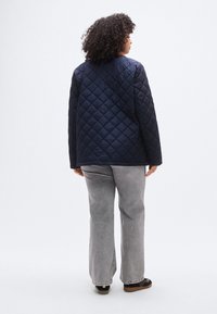 Μπλε ναυτικό καπιτονέ μπουφάν με ίσια γραμμή και μακριά μανίκια, με υφασμένο μοτίβο σε σχήμα διαμαντιού και στρογγυλεμένο τελείωμα, που φοριέται πάνω από γκρι παντελόνια.