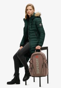 Giacca puffer verde con cappuccio foderato in pelliccia, leggings neri e stivali neri con lacci. Zaino rosa con logo "SUPERDRY" e cerniere.