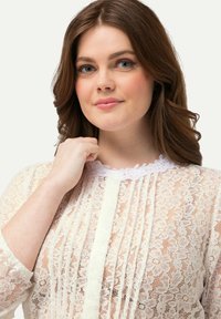 Blusa di pizzo color crema con motivi floreali e pieghe. Presenta un colletto bianco con bordo scallop e maniche lunghe. Materiale morbido e testurizzato.