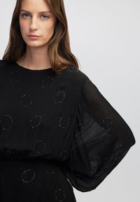 Schwarze Bluse aus durchsichtigem Stoff mit runden Perlenakzenten, weiten Ärmeln und Rundhalsausschnitt, die eine fließende Silhouette erzeugt.