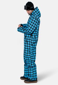 Mono de nieve de cuadros azules y negros con cremallera frontal, puños ajustables, capucha y diseño de ajuste holgado; se lleva con botas negras y gafas de protección.