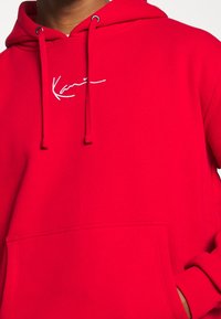 Roter Hoodie aus weichem Stoff, mit Kängurutasche und Kordelzügen. Weiße Stickerei mit dem Schriftzug "Kari" auf der Brust.