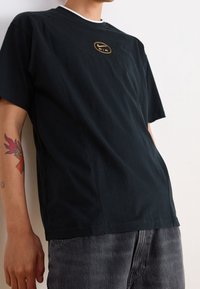 T-shirt noir à manches courtes en coton, avec un col rond, des bords blancs et un logo "Air" doré sur la poitrine.