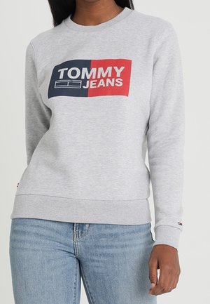 Kobieta ubrana w jasnoszary sweatshirt z granatowym i czerwonym logo "Tommy Jeans", zestawiony z jasnoniebieskimi dżinsami.