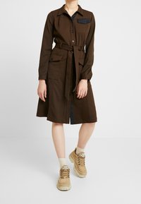 Ruskea paitamekko, jossa on pitkät hihat, napitus, kaksi etutaskua ja vyötäröllä vyö. Sisältää tummansinisen farkkutaskun. Yhdistetty beigen värisiin lenkkareihin.