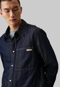Camicia in denim blu scuro con colletto a punta, chiusura frontale con bottoni e taschino con etichetta Calvin Klein. Texture liscia e design minimale.