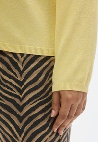 Rumeno pleteno pulover z rebrastim robom, kombiniran s krilom s črno-rjavim črtastim vzorcem. Gladka tekstura in prilegan silhueti sta očitna.