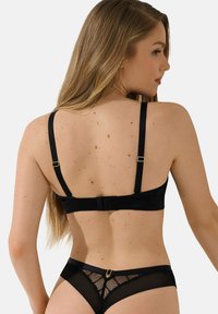 Ensemble de lingerie noir comprenant un bralet avec des bretelles réglables et une fermeture à crochets, associé à une culotte transparente avec un dos en mesh et des éléments de finition.