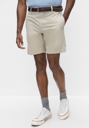 Mand iført beige shorts med brunt flettet bælte, grå sokker, hvide sneakers og lyseblå skjorte, stående mod en hvid baggrund.