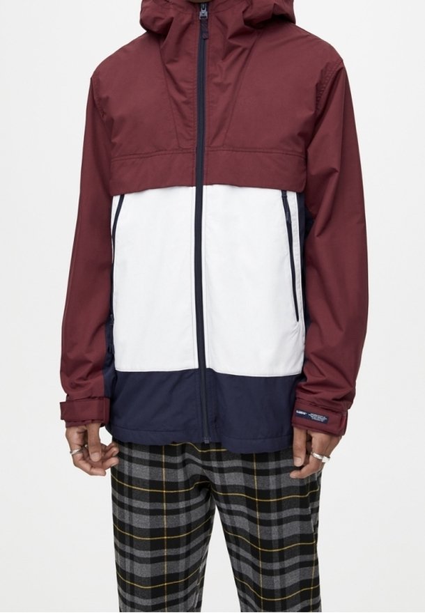 Color-block jas met een bordeauxrode capuchon, wit torso en donkerblauwe onderkant. Voorzien van een ritssluiting aan de voorkant en lange mouwen. Getextureerd, lichtgewicht materiaal.