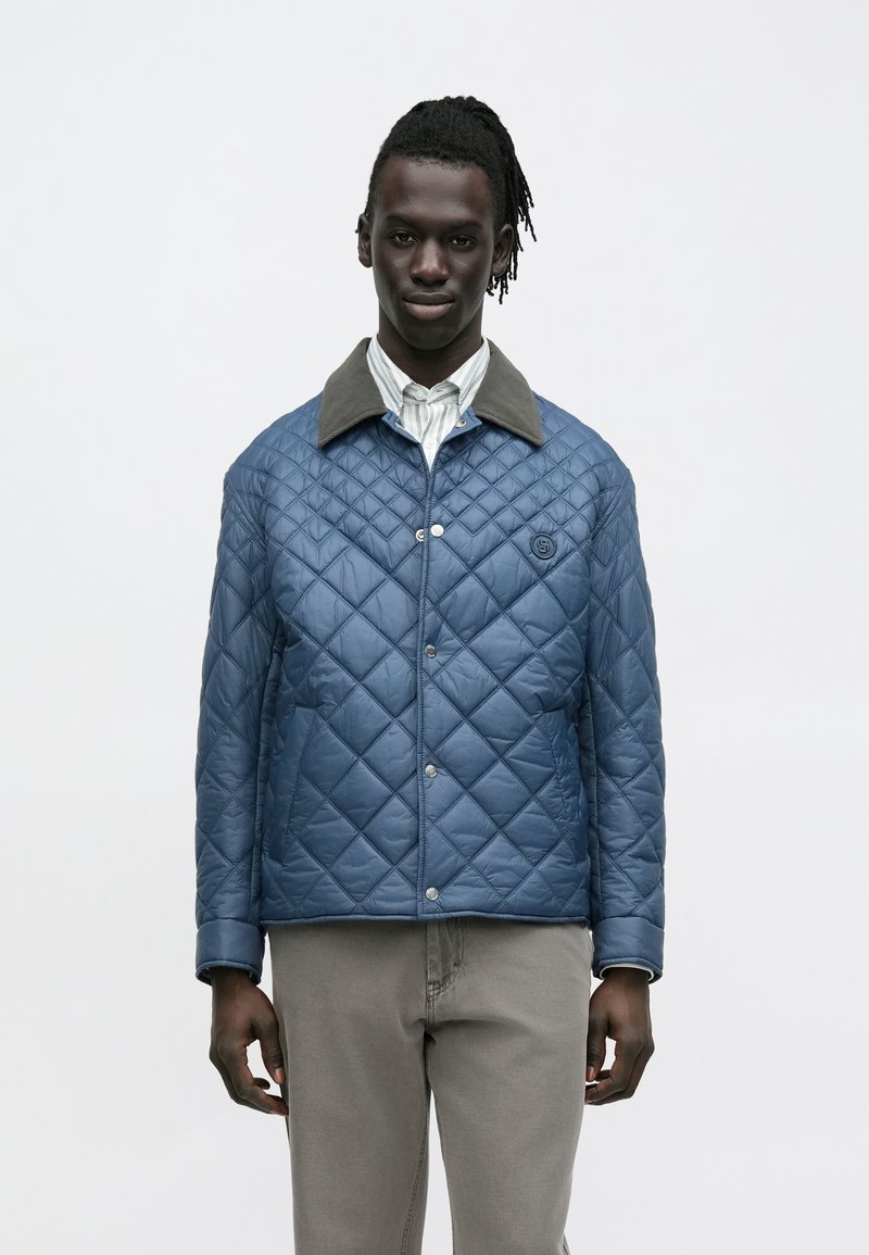 Gesteppte blaue Jacke mit einem grauen Kragen, ausgestattet mit einem Druckknopfverschluss und Seitentaschen. Glatte Textur und Steppmuster in Rautenform.