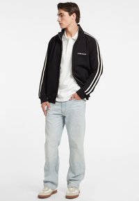 Giacca nera con zip e strisce laterali bianche, indossata sopra una polo bianca. Jeans azzurri chiari e sneaker beige completano l'outfit.