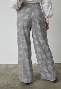 Grå rute brede bukser med en struktureret overflade, der har høj talje og pressede folder, set fra ryggen på en neutral baggrund.