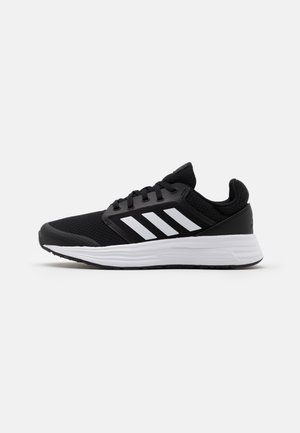 Zapatilla de running Adidas negra con rayas blancas y una suela acolchada blanca gruesa sobre un fondo claro y sencillo.