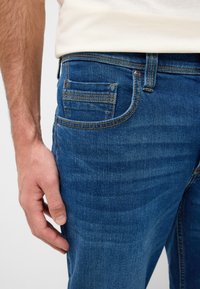 Pantalones vaqueros de denim azul oscuro con una vista cercana, que presentan un ligero desgaste, un bolsillo con detalles de costura y un acabado texturizado.