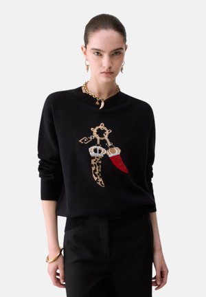 Donna con capelli scuri raccolti indietro che indossa un maglione nero con un design di pugnale oro e rosso, abbinato a pantaloni neri e gioielli dorati.
