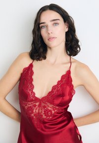 Rote Satin-Camisole mit Spitzenverzierungen am Oberteil und Saum, dünnen verstellbaren Trägern und einem V-Ausschnitt-Design. Glatte Textur.