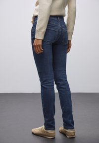 Jeans de mezclilla oscuros y ajustados con bolsillos traseros, combinados con un suéter claro y zapatillas beige, vistos de lado.