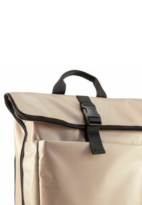 Beige waterdichte tas met een opvouwbare klep, zwarte rits en een voorvak. Beschikt over een handvat aan de bovenkant en een verstelbare schouderband. Gladde textuur.