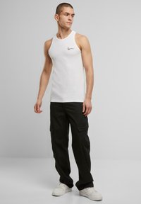 Weißes Baumwoll-Tanktop mit gerippter Textur, ausgestattet mit einem kleinen schwarzen Logo auf der Brust. Kombiniert mit lockeren schwarzen Cargo-Hosen und weißen Sneakers.