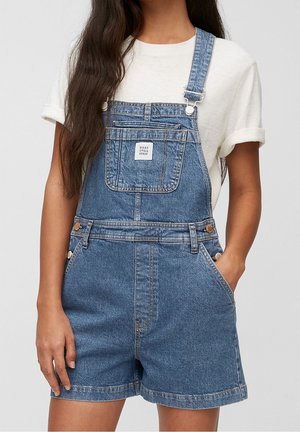 Femme portant une salopette courte en denim bleu avec une poche avant et un t-shirt blanc à manches retroussées, main dans la poche, sur un fond uni.