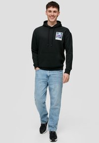 Sudadera negra con un bolsillo frontal y un parche gráfico; combinada con jeans de mezclilla azul claro y zapatillas negras. Diseño sencillo y ajuste casual.