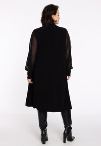 Robe noire avec des manches longues transparentes, une coupe décontractée et une longueur au niveau du genou, associée à des bottes noires à talons hauts et ajustées.