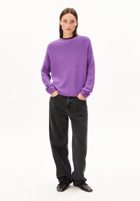 Junge Frau steht mit den Händen in den Taschen, trägt einen lockeren lila Pullover, weite schwarze Jeans und schwarze, Nieten besetzte Schuhe vor weißem Hintergrund.