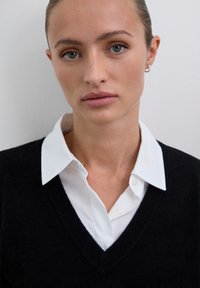 Junge Frau mit heller Haut und zurückgezogenem Haar, die ein weißes Hemd mit Kragen unter einem schwarzen V-Ausschnitt-Pullover trägt, vor einem schlichten hellen Hintergrund.