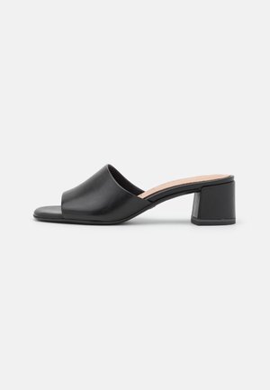Mule noire à bout ouvert avec large bride en cuir et talon carré sur fond blanc.