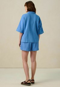 Chemise courte à manches courtes en tissu texturé bleu avec col et shorts assortis, offrant une coupe décontractée. Port é avec des sandales marron, vu de dos.