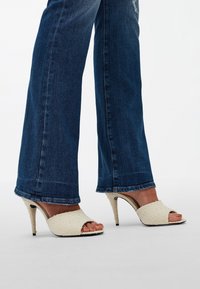 Escarpins beiges à bout ouvert, en tissu texturé, associés à un jean évasé bleu foncé. Les chaussures présentent un talon courbé et aucun accent décoratif.