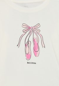 Rosa ballet-sko bundet med et bånd henger med ordet "BALLERINA" under et hvitt stoff bakgrunn.
