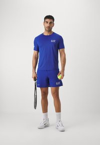 Conjunto atlético azul compuesto por una camiseta de manga corta y pantalones cortos, hecho de un tejido ligero, acompañado de zapatillas blancas y sosteniendo una raqueta de tenis y una bola.
