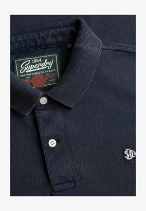 Marineblauw poloshirt met een gestructureerde stof, een kraag en twee witte knopen. Voorzien van een rood-groen merkembleem aan de nek.