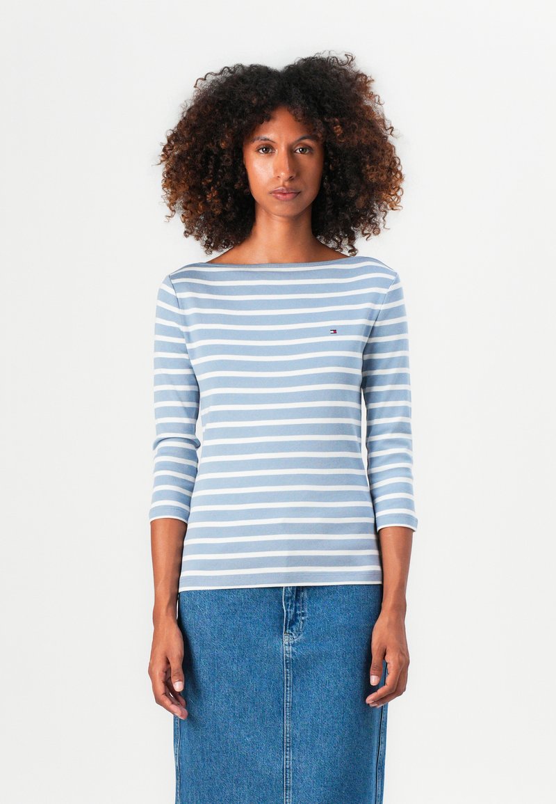 Stripet langermet topp med lyseblå og hvite horisontale striper, rund halslinning og logodetalj på brystet, kombinert med en denimskjørt.