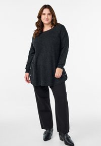 Črna tunika z dolgimi rokavi, izdelana iz teksturirane tkanine, z bočnimi gumbi, zaobljenim robom, v kombinaciji s črnimi širokimi hlačami in gležnjarji.